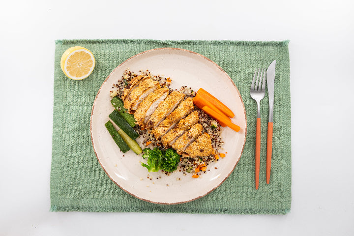 Plated dish with sliced chicken, vegetables, and a lemon wedge on a green placemat.