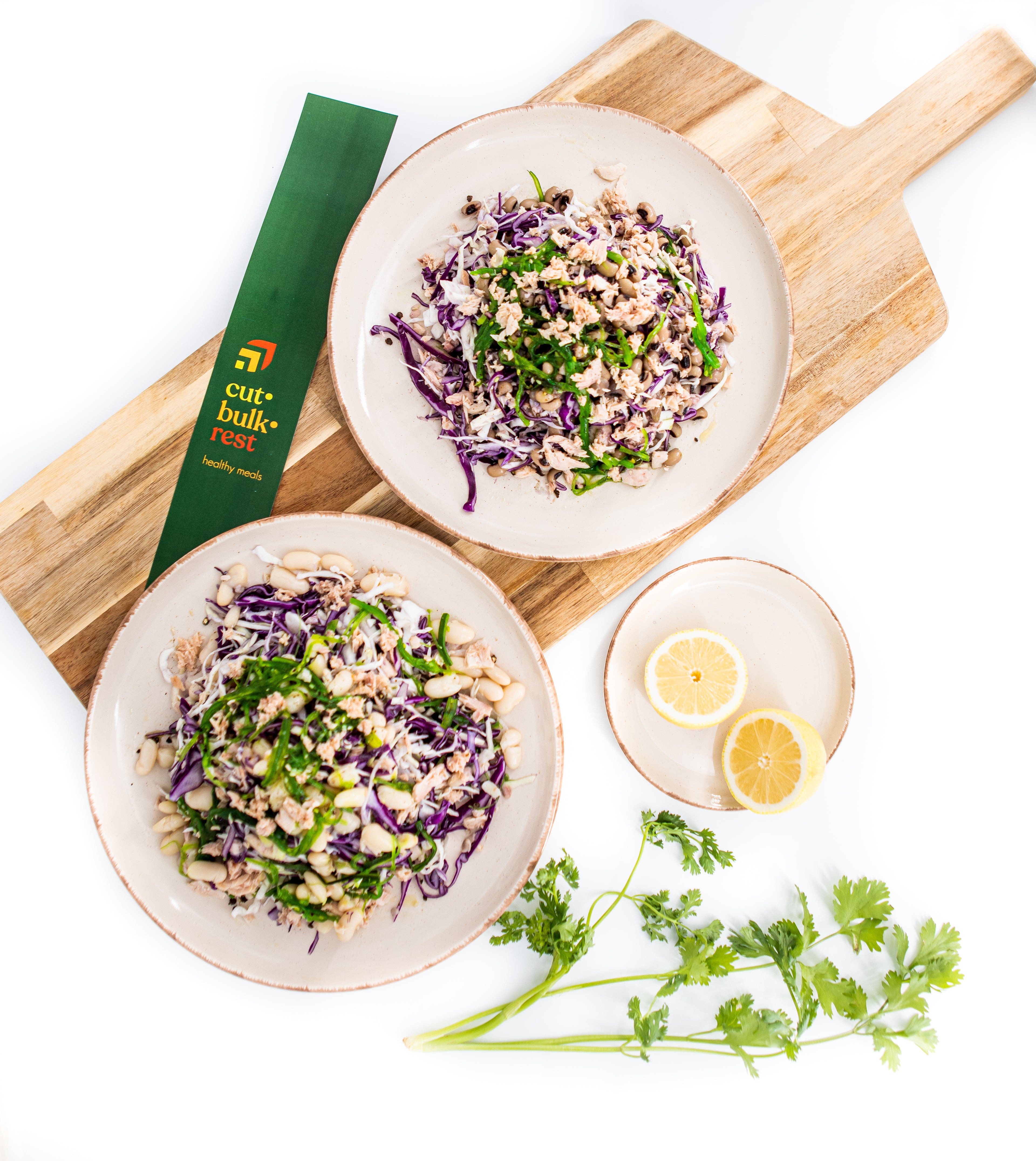 Two plates of salad on a wooden board with lemon wedges and herbs, featuring Curd Balkan brand.